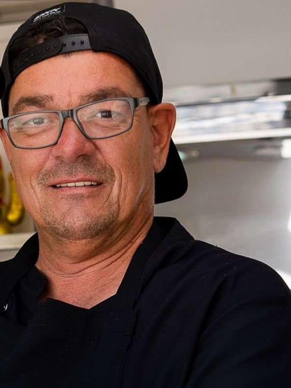 El cocinero de WURSTWERK con gorra negra y camiseta con el logo, sirviendo una salchicha en el truck
