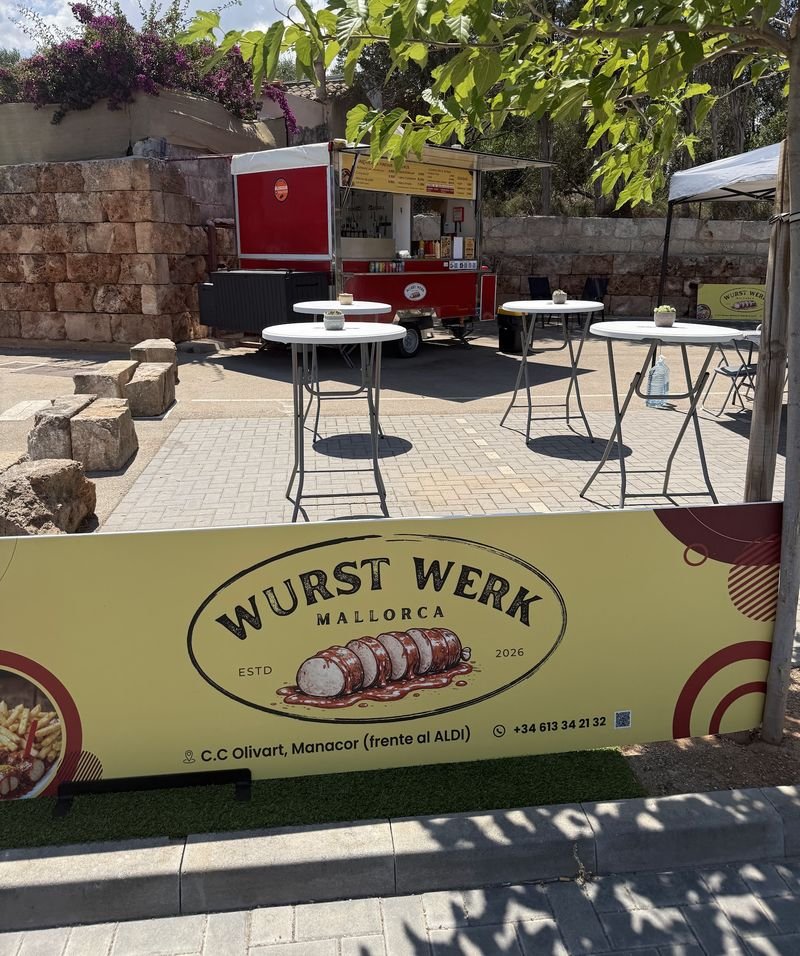 Mesas altas a la sombra junto al truck rojo de WURSTWERK, con la lona del logo en primer plano